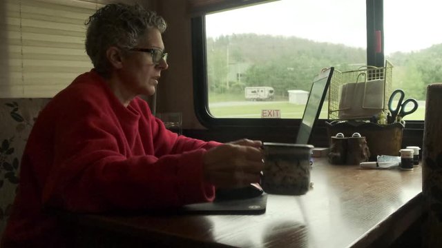 Caucasian woman works on laptop and drinks coffee inside fifth wheel RV camper, close up