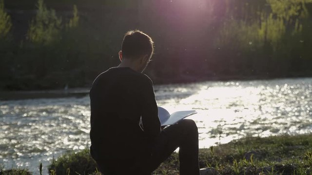 Close up of a young man sitting and reading a book close to the river in a big park in Florence, Italy. Peaceful moment, a break in the sunset