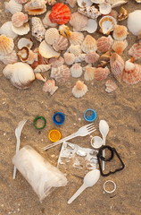 A lot of different plastic waste: disposable plastic cups,plastic bags, disposable spoons, forks, plastic lids, collected on the beach against the background of sand and beautiful shells. 