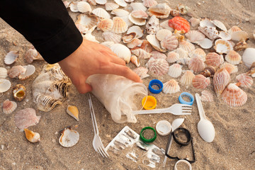 A man's hand collects and removes plastic waste on the beach: bags, disposable plastic appliances, plastic lids among the shells. 