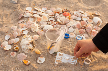 A man's hand collects and removes plastic waste on the beach: bags, disposable plastic appliances, plastic lids among the shells. The concept of ecology, cleaning the environment