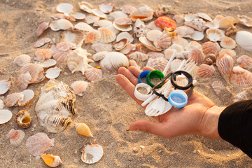 A man holds plastic lids and a disposable plastic fork and spoon in the palm of his hand against the background of beautiful seashells and sand on the beach. 