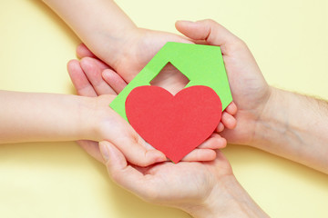 Obraz premium Close Up of dad and child hands holding green paper house with red heart on yellow background.