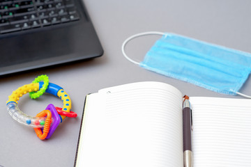 Protective medical mask near the laptop and pen with open notebook and rattle toy on gray background.