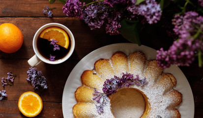 fresh orange muffin with orange slices and chocolate on a table with purple lilac flowers and a cup of black tea top view.