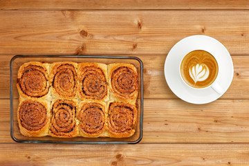 Homemade cinnamon rolls from yeast dough in transparent glass form and cup of coffee cappuccino on wooden table. Top view.