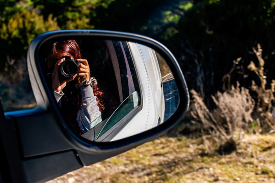 A close-up shot of the reflection of an unrecognizable young Caucasian redhead female photographer holding a DSLR photo camera in the rearview mirror of a moving car