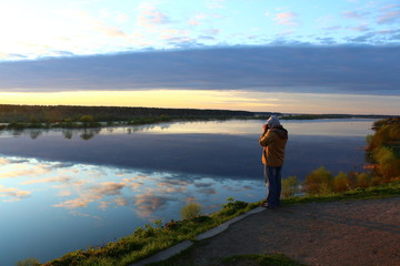 Fototapeta na wymiar A man in a jacket and hood illuminated at dawn with a camera on a high Bank takes a picture of Golden clouds shining in the river on a blue surface.Reflection of the sky in the water expanses.Russia