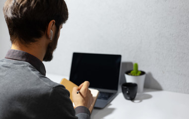 Back view of businessman working home, making notes in notebook, using wireless earphones, laptop, coffee mug and cactus on work desk. Background of textured wall of grey color.