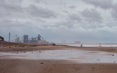 Woman and child walk along empty wet beach with heavy industrial steel plant in distance, in...