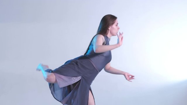 Medium long shot of young beautiful woman dancer in a dress with long hair emotionally dancing contemporary, modern ballet dance in the studio in color back light, slow motion, isolated