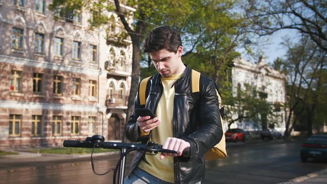Poartrait of delivery man with yellow bagpack standing with electric scooter in city and using smartphone to find the client's adress, slow motion