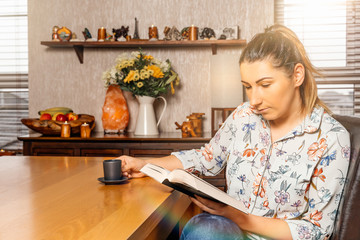 Young woman reading a book and drinking espresso from comfort of her home at sunny day during covid 19 or coronavirus pandemics and quarantine