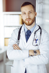 Red-bearded doctor standing straight in sunny clinic near his working place. Portrait of physician. Medicine concept