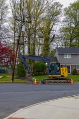 Yellow heavy equipment excavator parked on the side of a residential street in front of a house with orange warning cones in front and back
