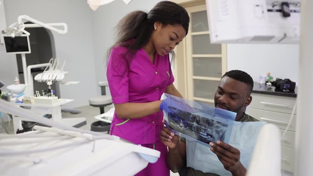 African male patient getting dental treatment in dental clinic. Pretty young African female dentist analyzing x-ray image, teeth photograph and explaining results to patient, man looking attentively