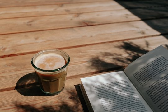 Iced Cappuccino Coffee in a recycled glass cup with vegan soy milk on a wooden floor near an open book in shade on a sunny day - Powered by Adobe