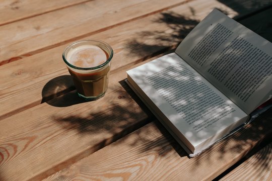 Iced Cappuccino Coffee in a recycled glass cup with vegan soy milk on a wooden floor near an open book in shade on a sunny day