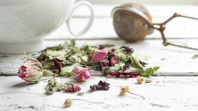 Herbal tea - lemon balm, linden, verbena, rosebuds, lavender, a white cup and tea infuser spoon on a light wooden table, close-up