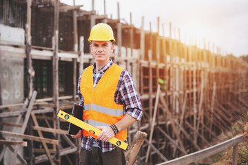 Fototapeta na wymiar Young caucasian man architect smiling and holding laptop with blueprint on a building construction site