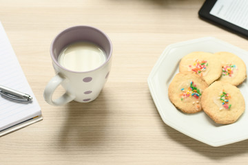 Vista de uma xícara com leite e biscoitos simples com confeitos coloridos, ao lado um leitor de ebook sobre um caderno.