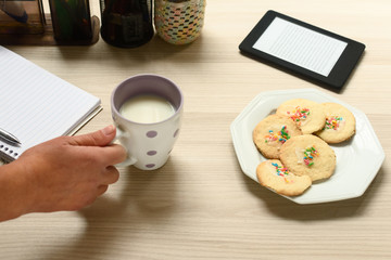 Mão segurando uma xícara com leite ao lado biscoitos simples com confeitos coloridos, cercados por materias de estudo, como caderno, caneta e um leitor de ebook sobre a mesa.