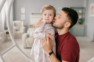 A handsome strong man in a bright room with his cute little daughter sit on a round swing and enjoy the pastime