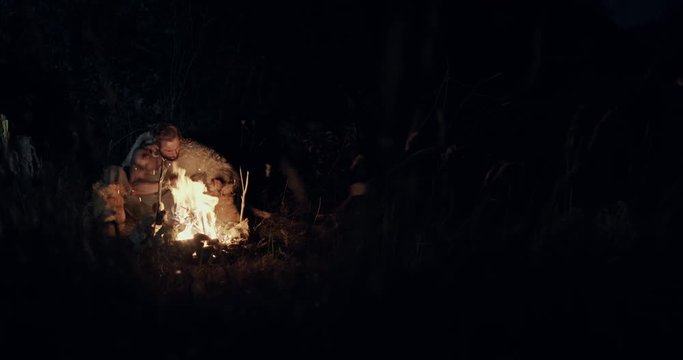 Male barbarian sitting near burning fire and sharpening knife on campsite at night in wilderness.