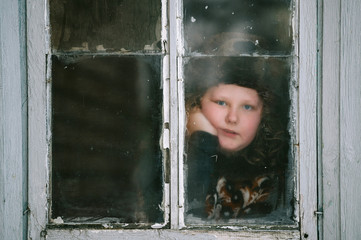 Portrait of a little girl with tranquil face expression looking out of old dirty window with reflections.