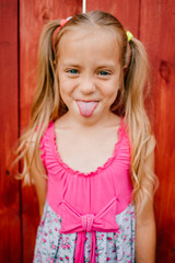Little cute girl with blond hair and pink dress makes faces against the wooden wall