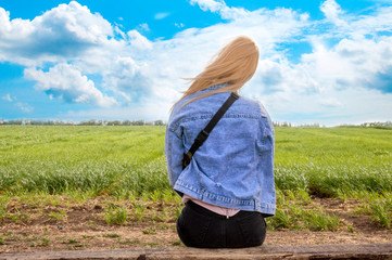 A young girl sits on a bench and looks at the field. Beautiful blue sky. Background green field and sky. The blonde is sad alone sitting on the bench. Beautiful girl admires nature.