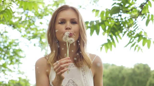 Beautiful attractive girl in a good mood in a white dress in the park admires and blows on a dandelion. Slow motion. Green around.