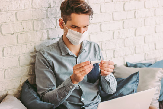 Sick businessman freelancer in medical face mask hold electronic thermometer in hand, work with laptop at home. Hipster man student in protective mask measure body temperature with digital thermometer - Powered by Adobe