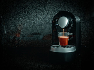Transparent glass cup of coffee on top of an espresso machine on a black and gray background, coffee cup full of creamy coffee