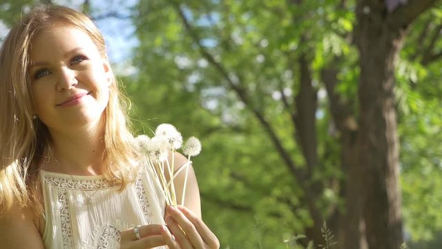 Beautiful attractive girl in a good mood in a white dress in the park admires and blows on a dandelion. Slow motion. Green around.
