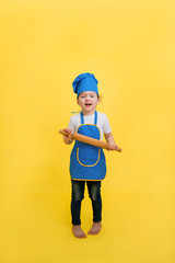 A little girl cook with a rolling pin on a yellow background with an empty seat. A beautiful girl in a full-length apron and hat.