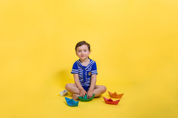 A beautiful girl in a sailor costume sits and holds a green paper boat in her hands. Free space on a yellow background. The day of the Navy.