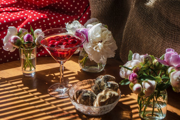 Solemn rustic still life with glass of red drink with cakes in a bowl surrounded by white flowers on textile background on wooden table. Diagonal striped shadow of the blinds on bright sunny day