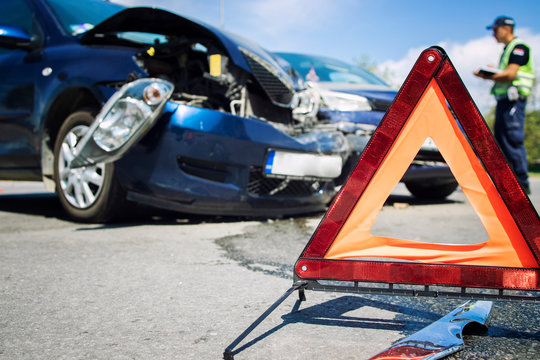 Car accident. Road disaster. Vehicle collision. Warning triangle for traffic or damaged cars in the middle of an intersection. In background police officer writing an accident report. Car insurance. c