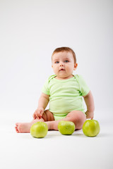 Cute toddler in green bodysuit sits with green apples on white background