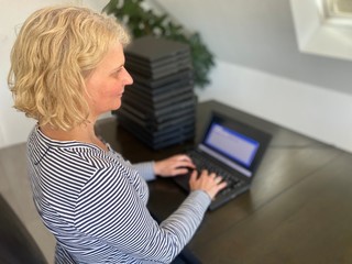 Corona Crisis: A casually dressed caucasian middle aged woman is installing a laptop computer with a stack of several more computers next to her. Laptops are used and second hand, might be for people