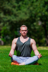 Naklejka na ściany i meble Young muscular man is doing yoga in a summer park. Sporty man is sitting in the Lotus position and relaxing outdoors