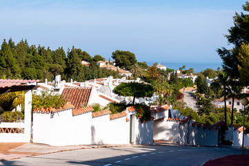A winding road in a spanish village among white houses with red roofs and green trees leading down to the sea.
