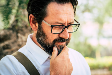 Worried adult caucasian 50 years old man touch the beard and think alone - eyeglasses and people outdoor with green park defocused background