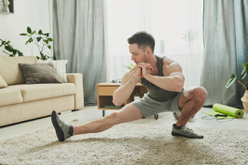 Young active man in sportswear doing stretching exercise for legs on the carpet