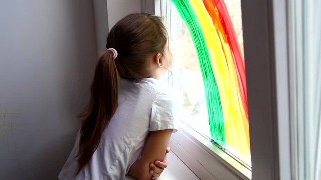 A little girl looks out the window with a painted rainbow during coronavirus quarantineself-isolation at home. Chase the rainbow flashmob and staying at home concept.