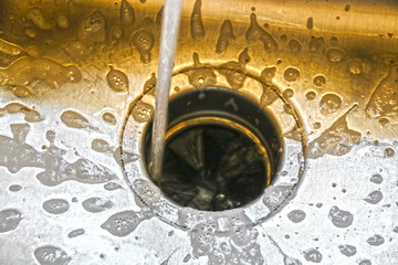 Water running into a garbage disposal of a stainless steel sink with gold toned blurred background...