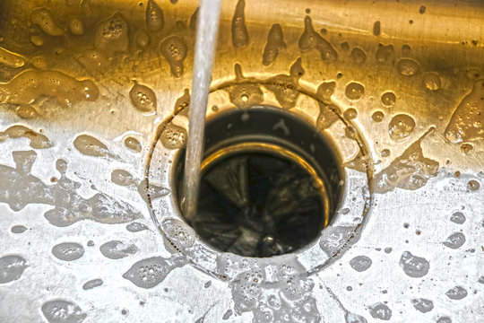Water running into a garbage disposal of a stainless steel sink with gold toned blurred background and sharp silver foreground with soap bubbles draining toward hole