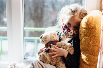 A sick child wearing a protective mask sits on the window. Patient isolated in house to prevent infection. Coronavirus. Teaching your child preventive measures against covid-19, viruses and flu. 