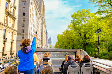 Tourists enjoying views of New York City from top of open roof bus in spring; woman in blue is...